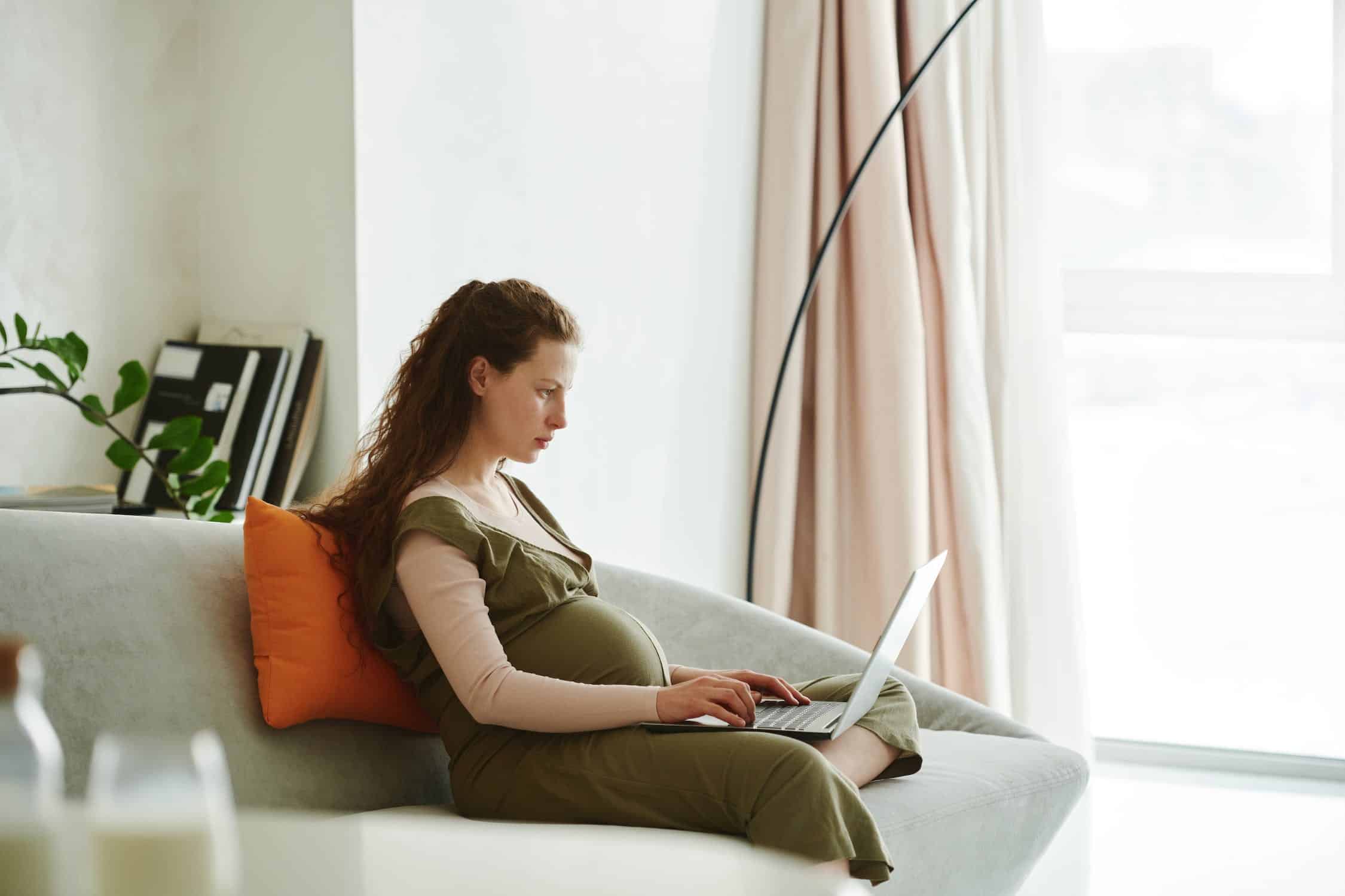 pregnant woman in a green dress sitting on a white sofa looking at a laptop learning about maternity rights