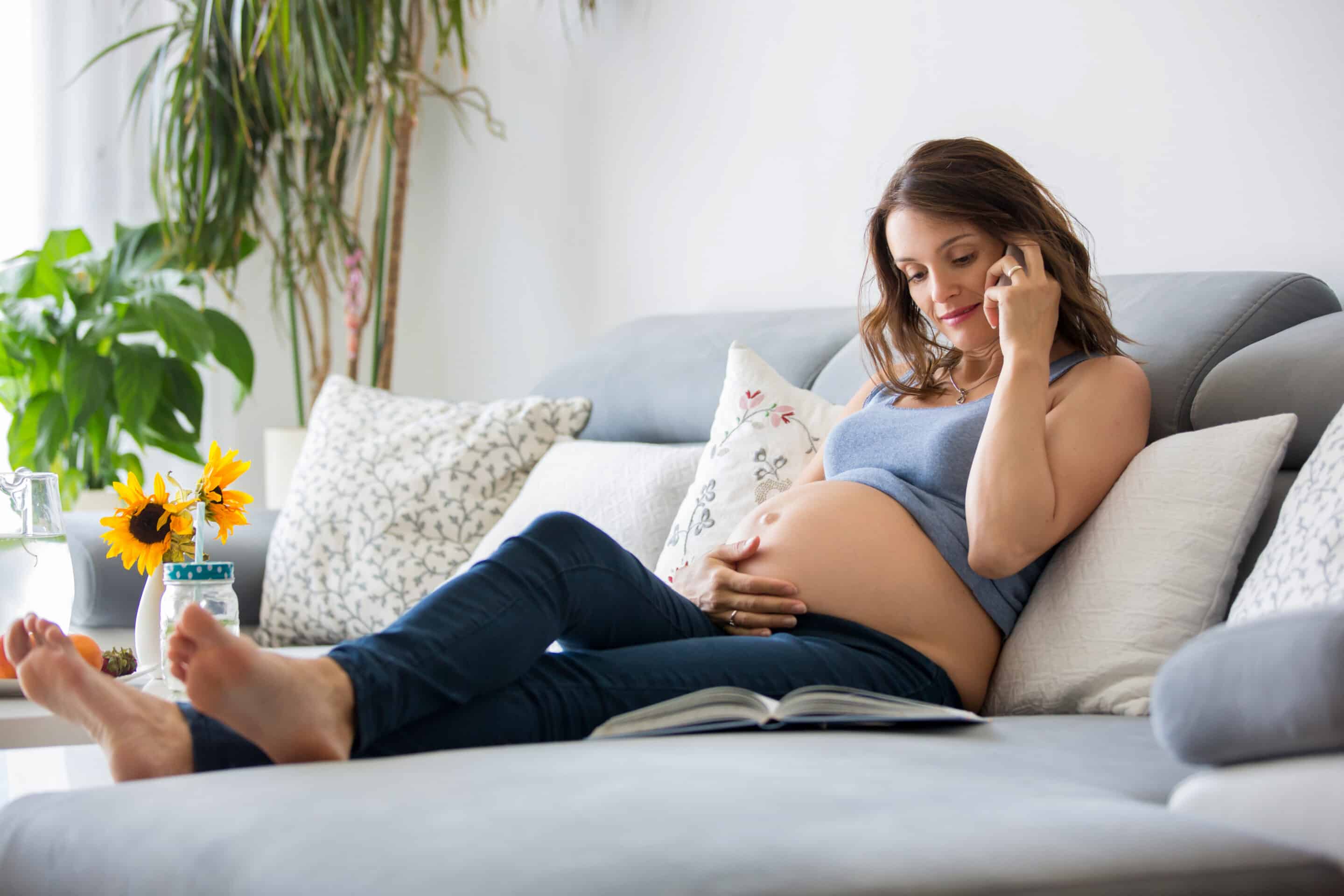 Pregnant woman sitting on the sofa talking on the phone with an open book. Taking care of mental health