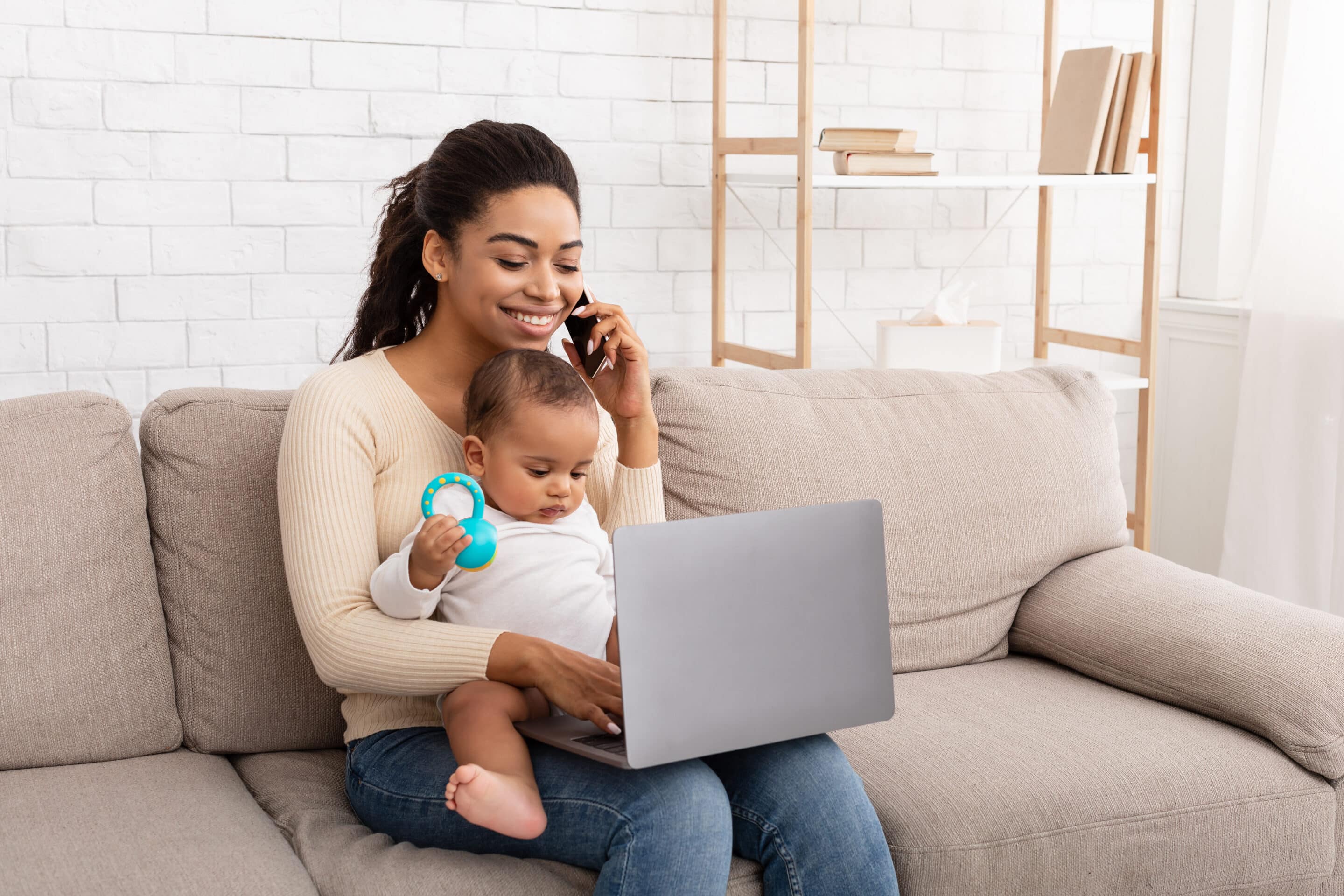 A mother sits with her baby on her lap whilst typing on a laptop and talking on the phone.